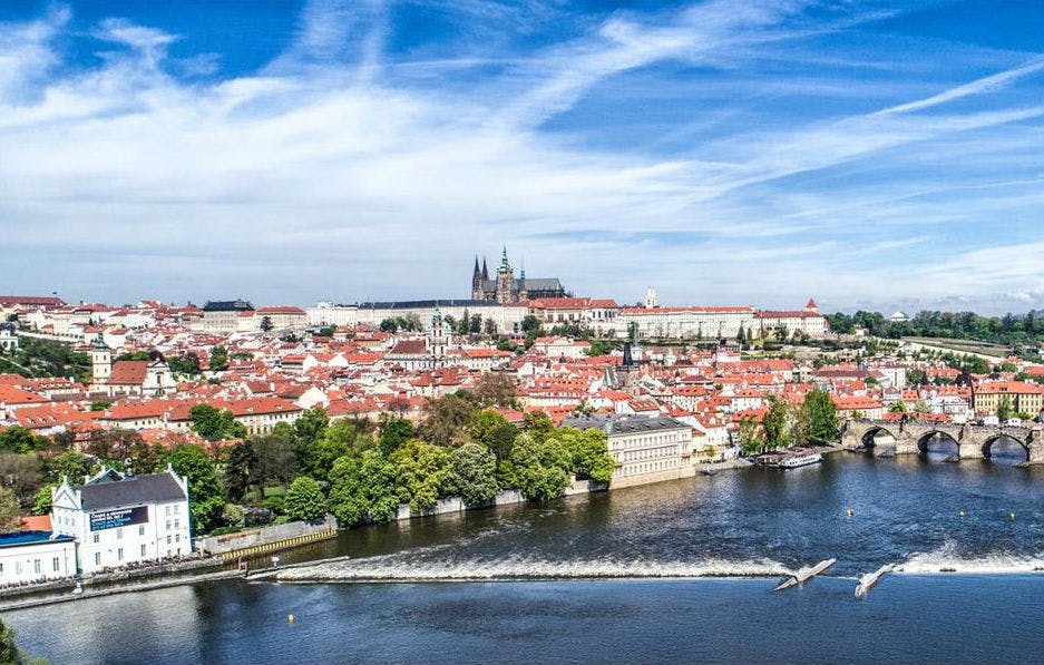 구름이 흩어져 있는 푸른 하늘 아래, 역사적인 건물과 강가의 붉은 지붕이 있는 도시의 파노라마 전망.