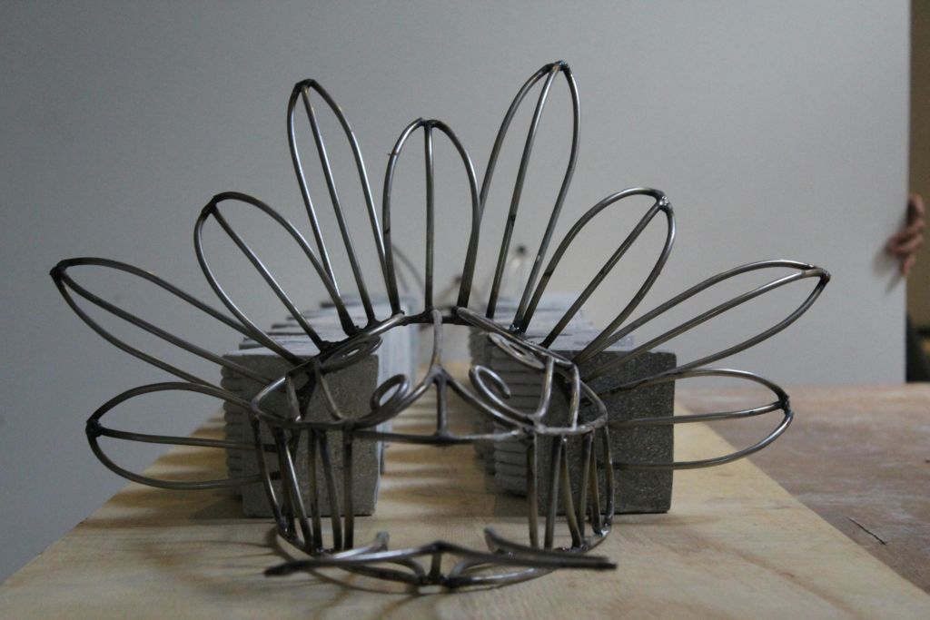 중립 배경에 꽃잎 모양의 고리가 있는 왕관을 닮은 와이어 조각. 콘크리트 블록이 구조물을 지지합니다.