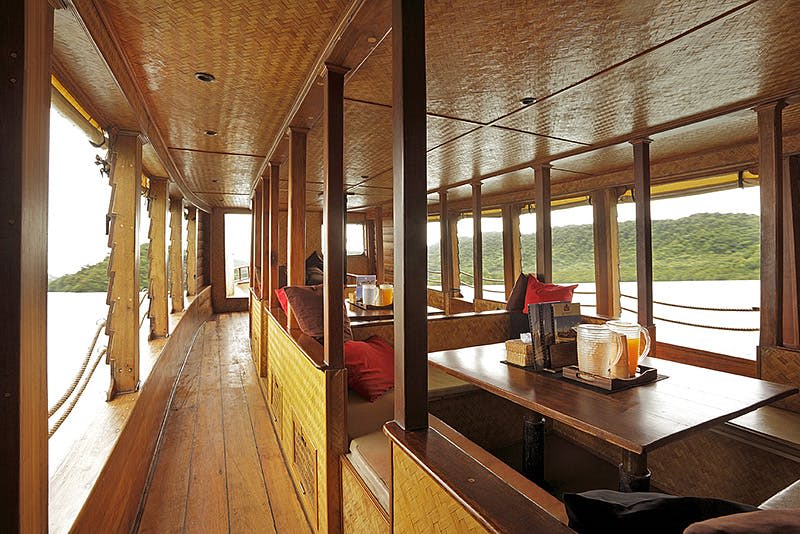 우븐 장식, 쿠션이 있는 좌석, 테이블, 창문을 통해 물과 녹지의 경치를 감상할 수 있는 나무 보트의 내부.