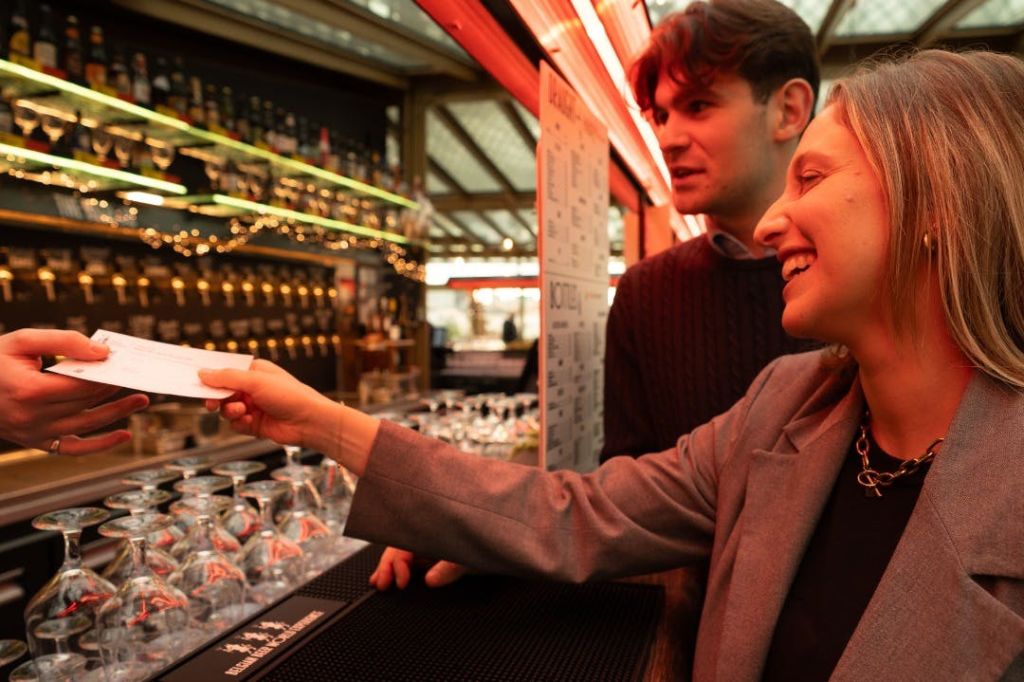 유리와 목재 장식으로 따뜻하게 빛나는 실내 환경에서 두 사람이 카운터에서 미소 짓고 상호 작용하고 있습니다.