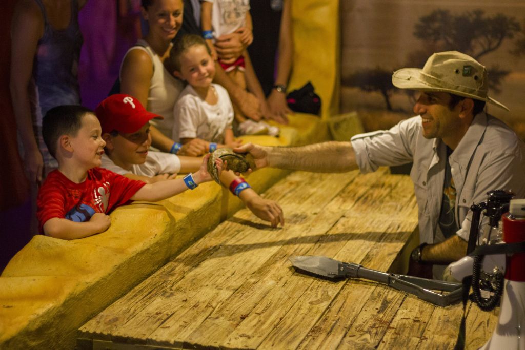 카우보이 모자를 쓴 남성이 나무 장벽 너머로 미소를 짓는 아이들에게 작은 동물을 보여주고 있습니다.