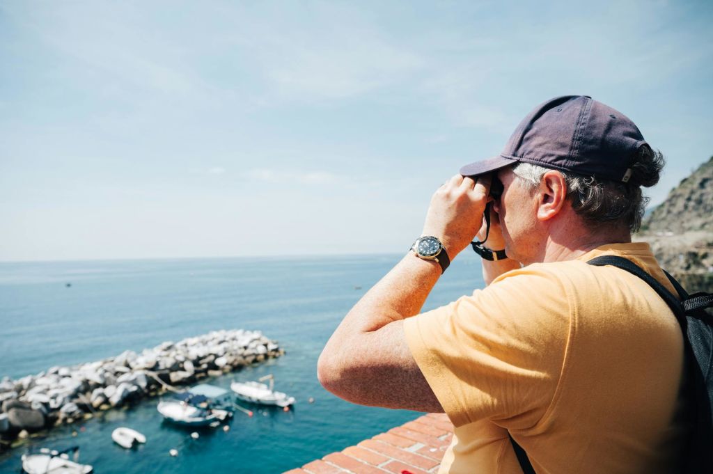 벽돌 난간이 있는 발코니에서 쌍안경을 사용하여 노란색 셔츠와 모자를 쓴 사람이 바다를 바라보고 있습니다.