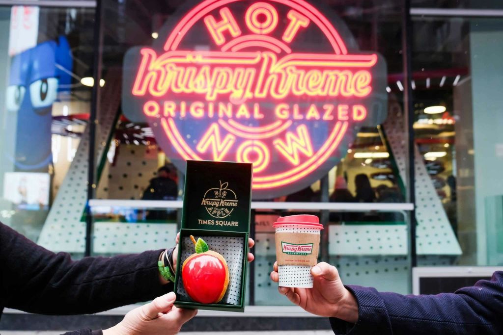 네온 "Krispy Kreme Original Glazed Hot Now" 표지판 앞에서 두 손으로 도넛 상자와 커피잔을 들고 있습니다.