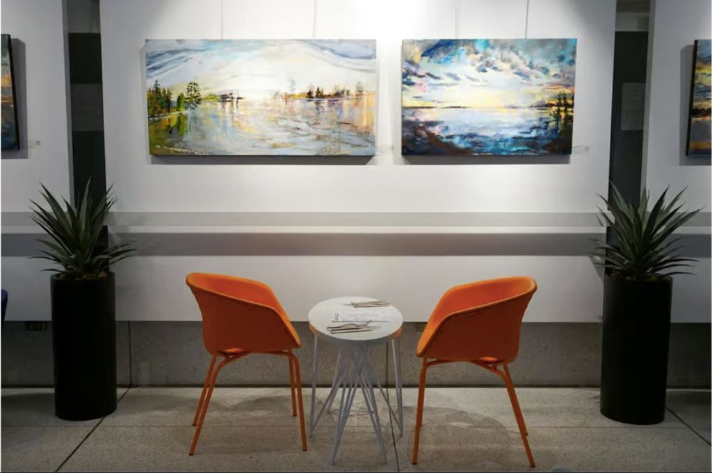 밝은 실내 조명 아래 두 개의 풍경화를 전시한 벽 앞에 주황색 의자 두 개가 놓인 작은 테이블.