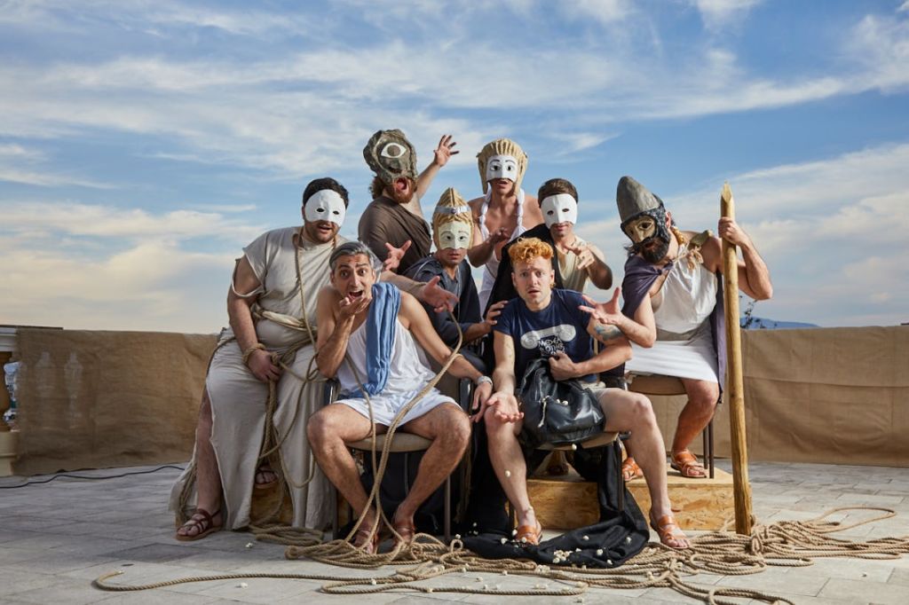 푸른 하늘을 배경으로 다양한 가면과 의상을 입은 사람들이 무대에서 극적인 포즈를 취하고 있다.