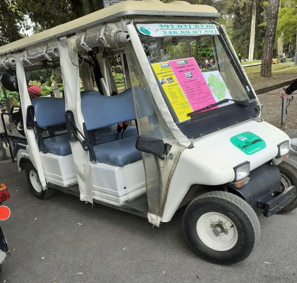 앞유리에 파란색 좌석과 광고가 있는 흰색 전기 카트가 공원 설정의 포장 도로에 주차되어 있습니다.