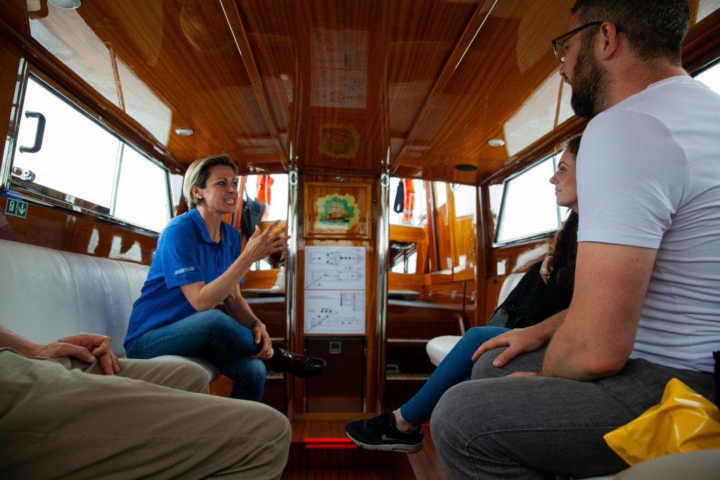 파란색 셔츠를 입은 여성이 나무 보트 안에 앉아 음료를 들고 있으며, 주변에는 다른 두 사람이 부분적으로 보입니다.