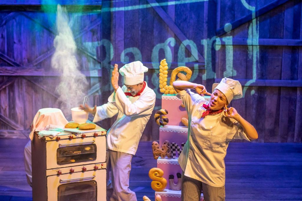 흰 유니폼과 모자를 입은 두 명의 셰프가 무대에서 공연합니다. 한 명은 얼굴을 가리고, 다른 한 명은 빵 소품으로 귀를 가립니다.