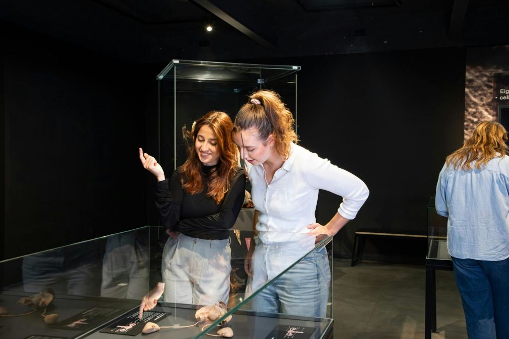 조명이 어두운 전시 공간 내부에서 유리 진열 케이스에 담긴 물건을 살펴보는 두 여성.