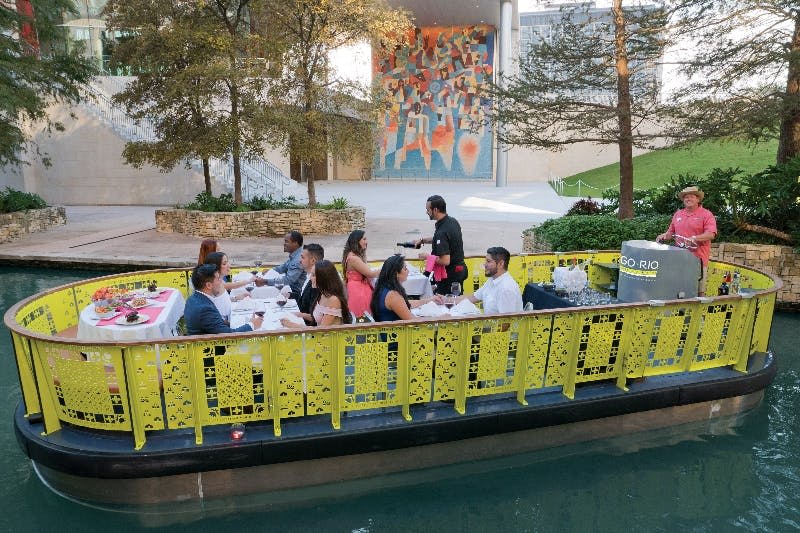 투어 가이드와 함께 노란색 리버보트를 타고 식사를 즐기는 사람들. 배경에는 나무, 화려한 벽화, 현대적인 건물이 포함됩니다.