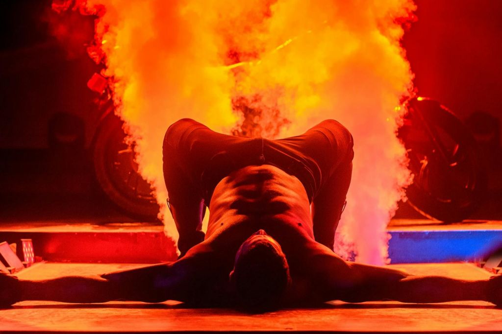 셔츠를 벗은 남성이 무릎을 구부린 채 등을 대고 누워 활기찬 주황색 연기를 내뿜으며 드라마틱한 장면을 연출합니다.