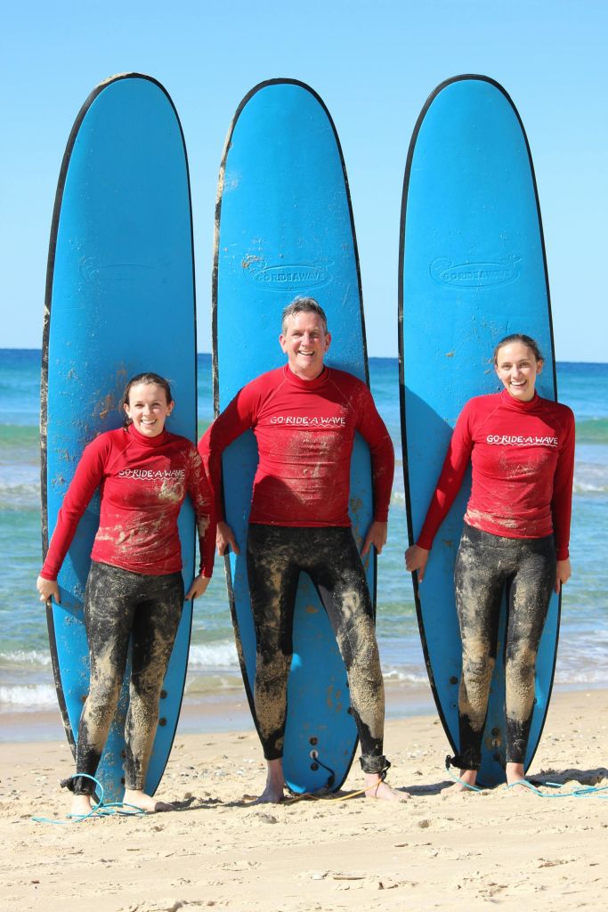 바다와 푸른 하늘을 배경으로 해변에서 파란색 서핑 보드를 들고 있는 빨간 셔츠를 입은 세 사람.
