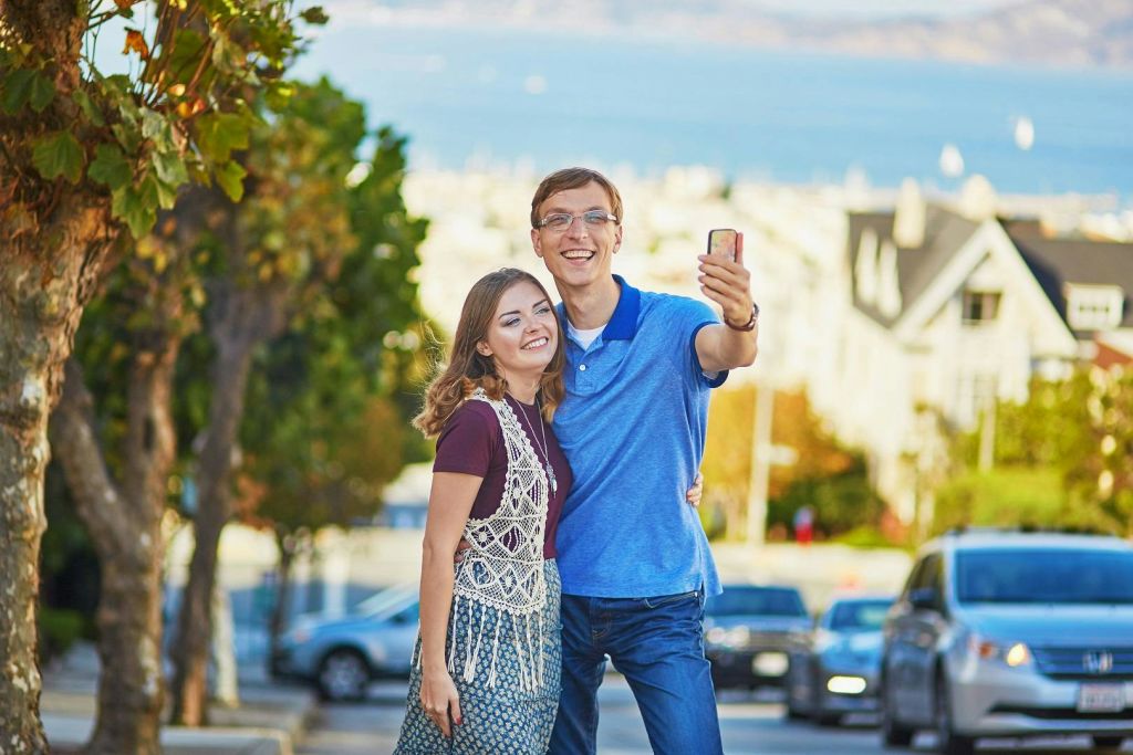 웃고 있는 커플이 거리에 서서 셀카를 찍고 있습니다. 배경에는 나무와 건물이 보이고, 물의 풍경을 감상할 수 있습니다.