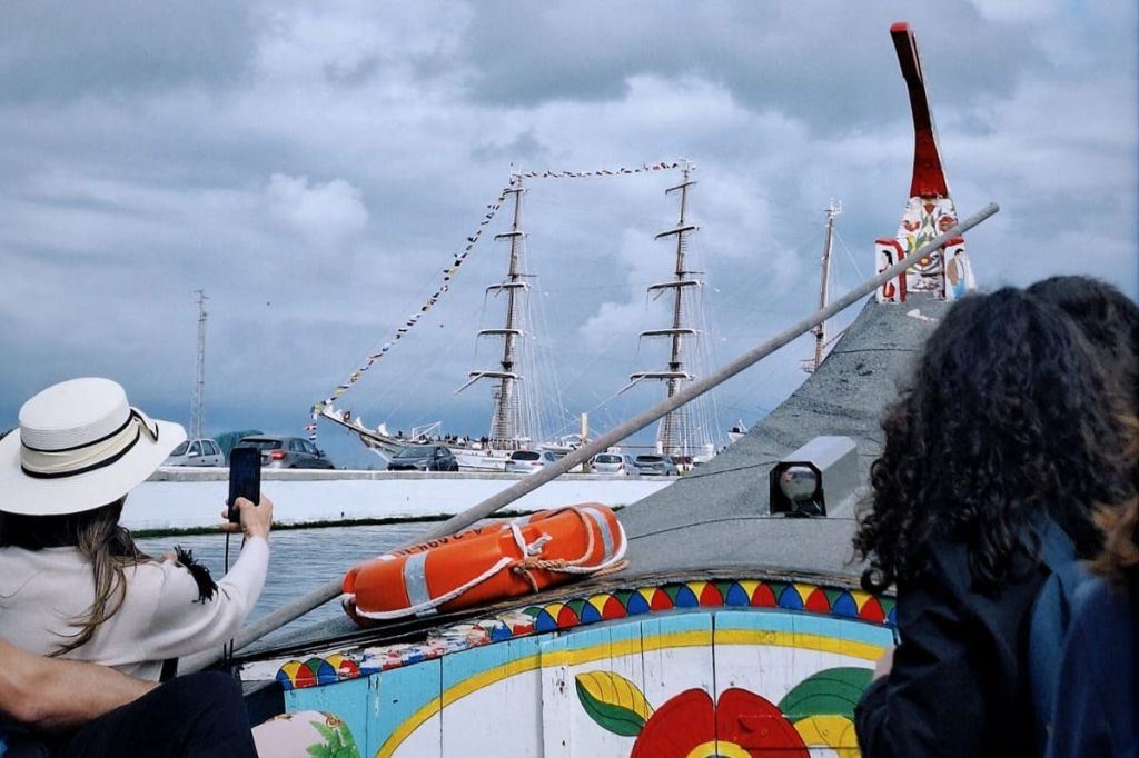 한 사람이 도킹한 높은 배에 장식용 깃발이 달린 사진을 찍고 있습니다. 전경에는 다채로운 색상의 보트가 있고 옆에는 구명보이가 있습니다.