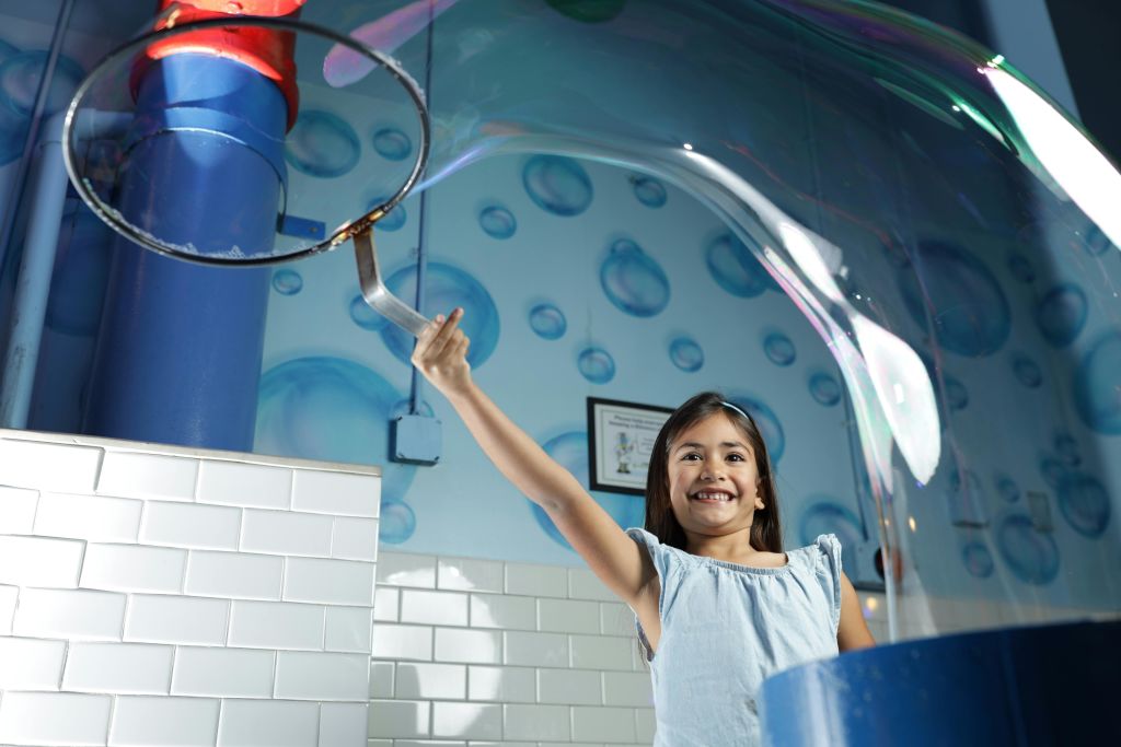 연한 파란색 드레스를 입은 한 소녀가 거품을 테마로 한 배경과 흰색 타일 벽이 있는 거대한 거품 안에 서서 미소를 짓고 있습니다.