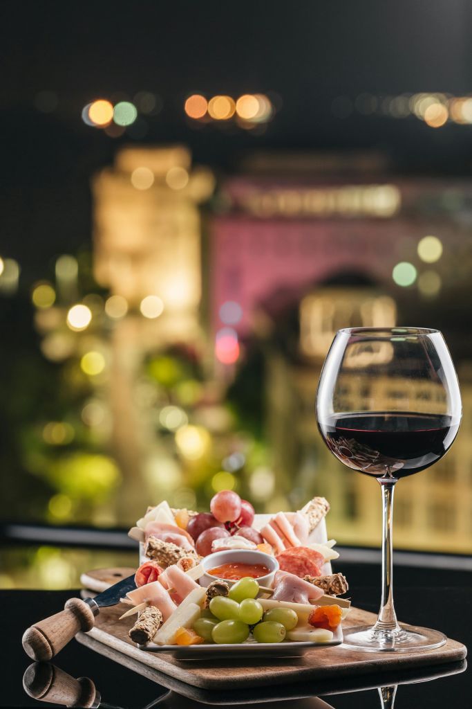 밤에 흐릿한 도시 풍경 앞에 고기, 치즈, 포도, 크래커를 곁들인 레드 와인 한 잔과 샤퀴트리 보드.