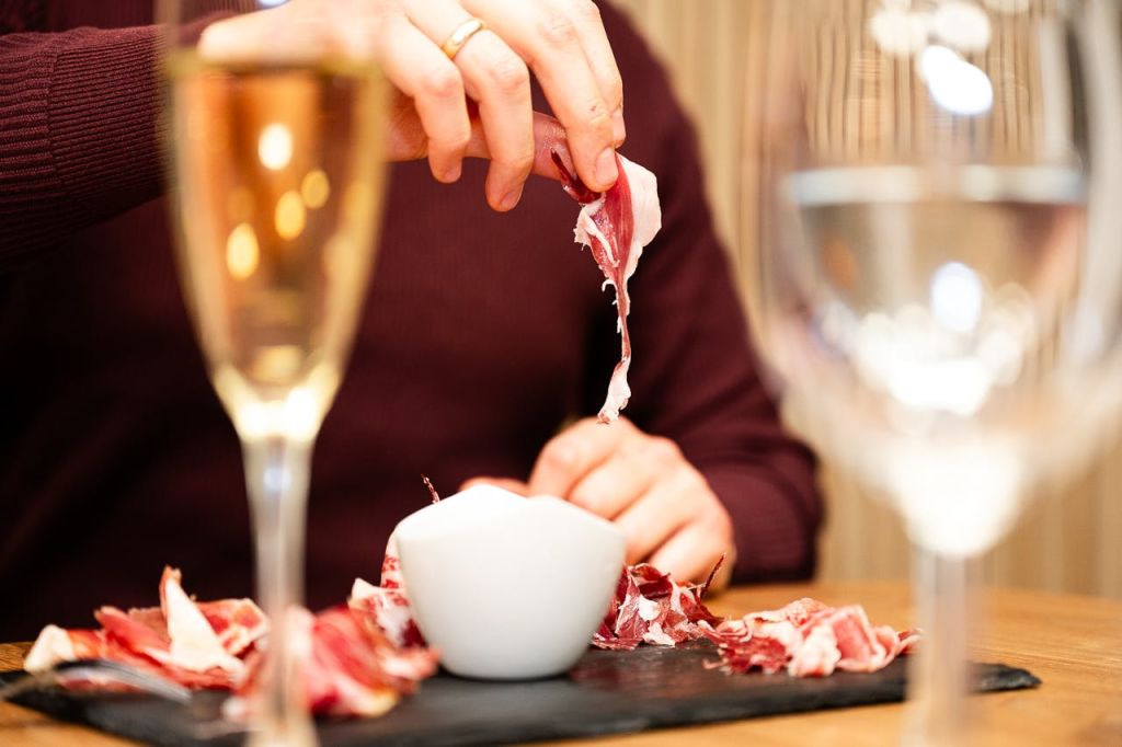 더 많은 고기와 작은 흰색 그릇이 담긴 접시 위에 잘게 썬 고기 조각을 들고 근처에 샴페인 한 잔을 들고 있는 사람.