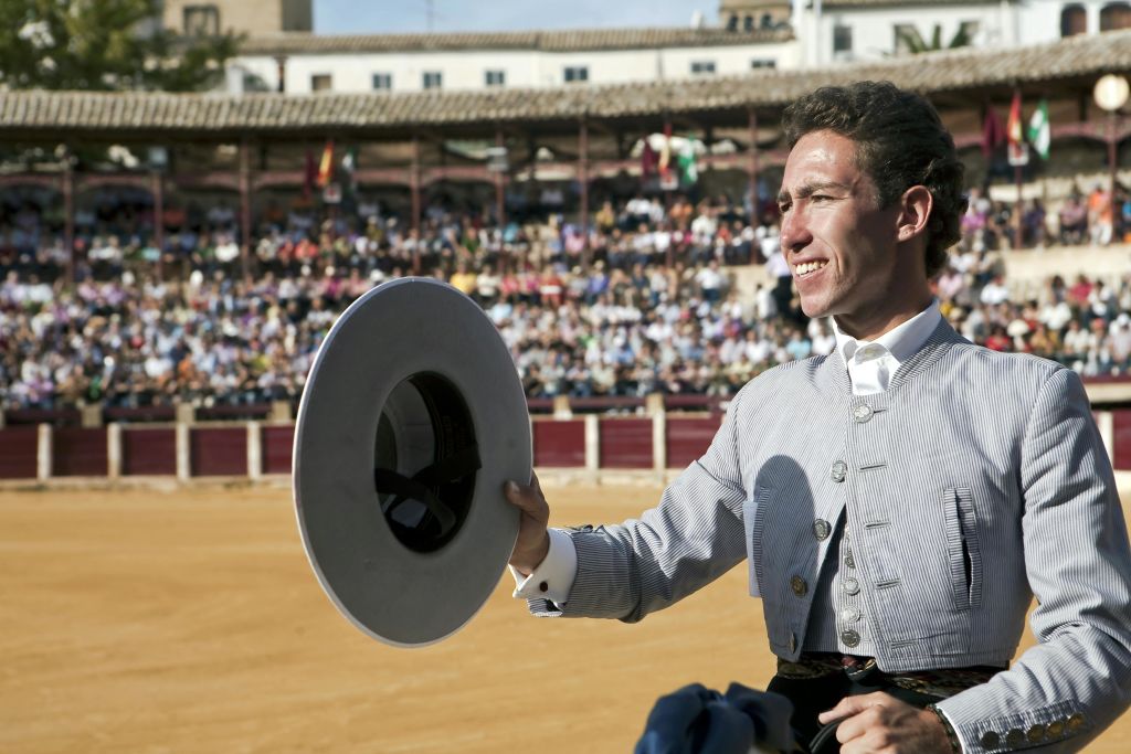 전통 복장을 한 남성이 모자를 쓰고 미소를 짓고 있으며, 군중은 황소 고리에 앉아 있습니다.