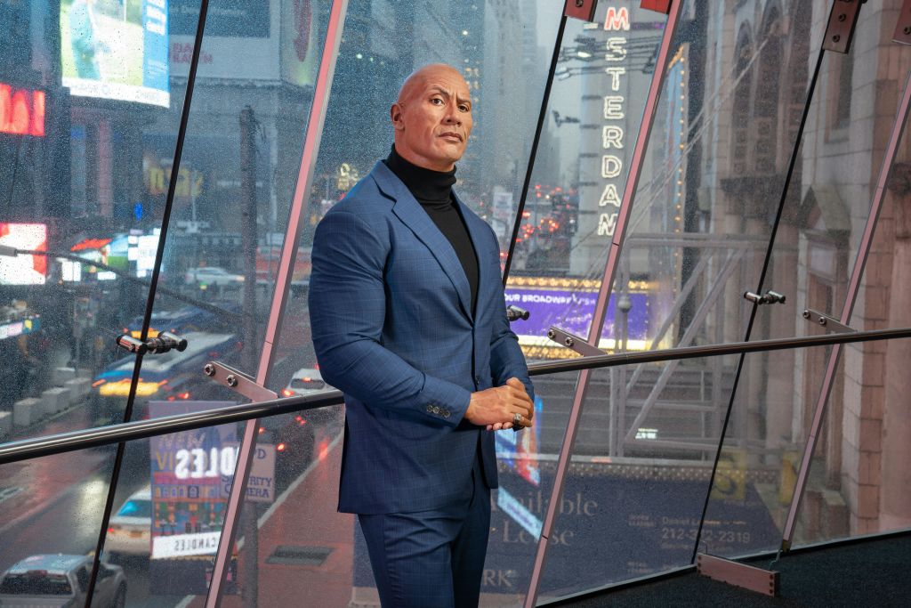 파란색 정장을 입은 남성이 네온사인과 도시의 불빛을 배경으로 커다란 창문 옆에 서 있습니다.