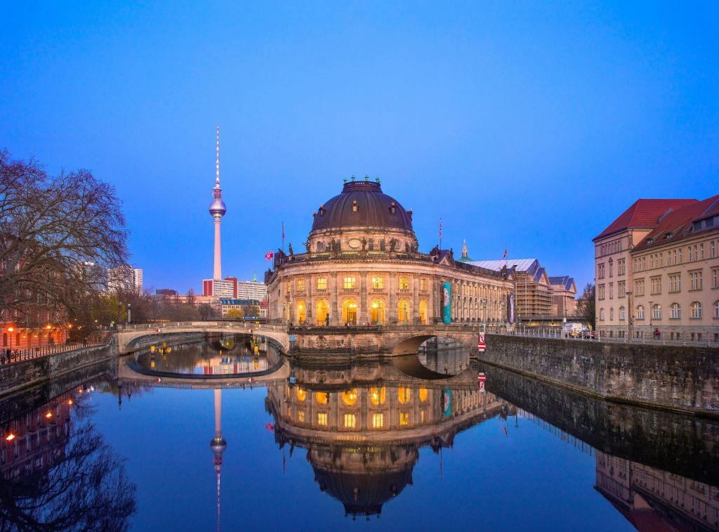 밝은 창문이 있는 돔형 건물이 강에 반사되어 맑은 하늘 아래 배경에 타워와 다리가 보입니다.
