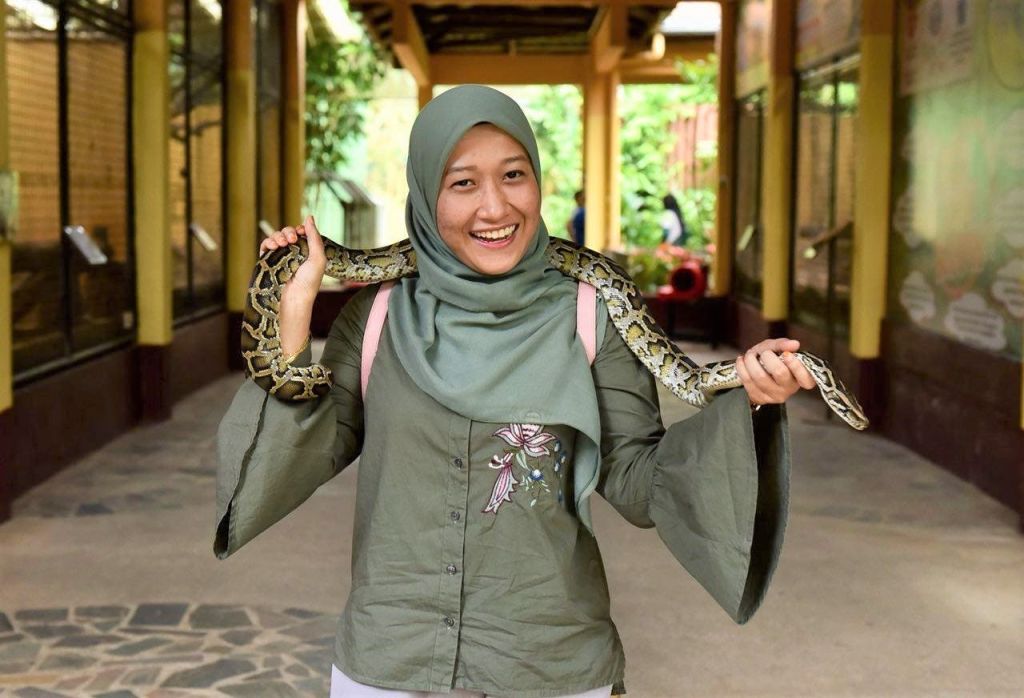 녹음이 우거진 야외 공간에 서서 뱀을 어깨에 두르고 미소를 짓는 여성.