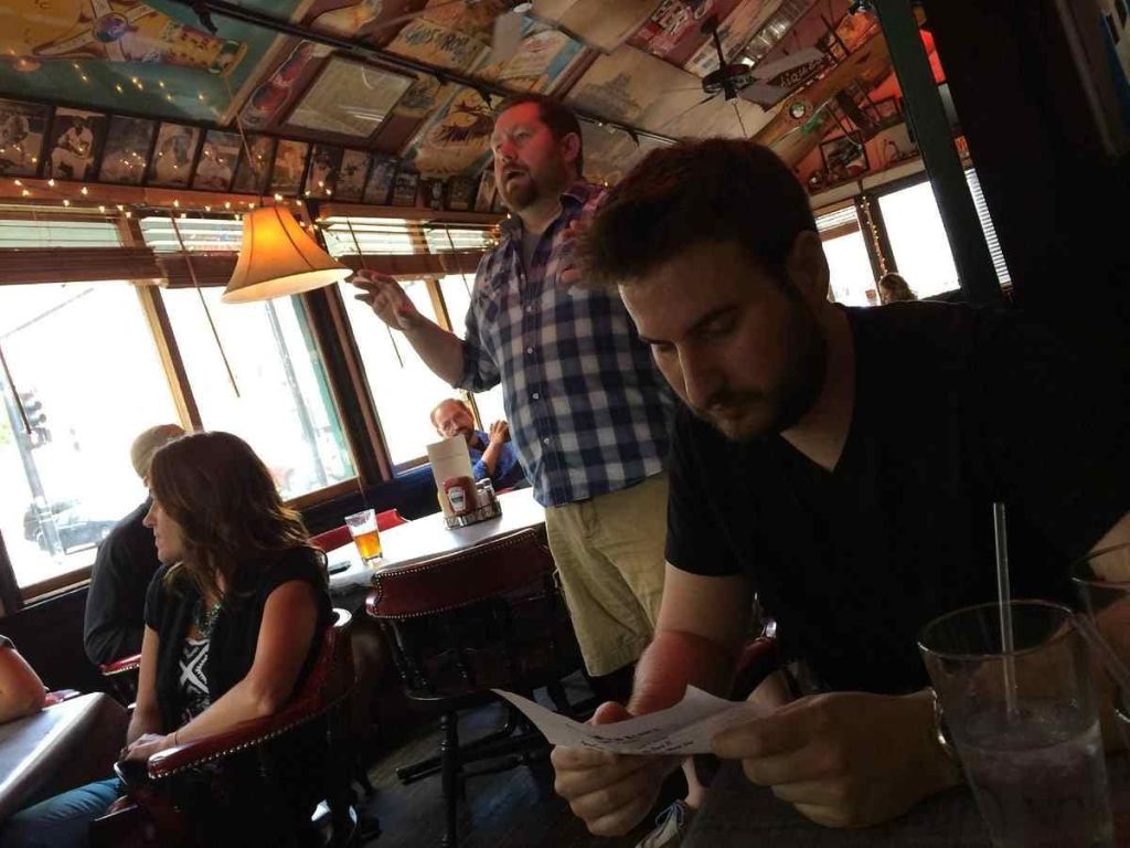 한 남자가 테이블에서 종이를 읽는 동안 다른 남자가 희미한 조명의 레스토랑에 앉아 몇 명의 다른 앉아있는 후원자들과 함께 제스처를 취합니다.