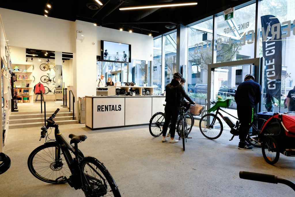'대여소' 카운터가 있는 자전거 매장의 사람들, 전시된 자전거, 커다란 창문, 카운터 뒤에 직원이 있습니다.