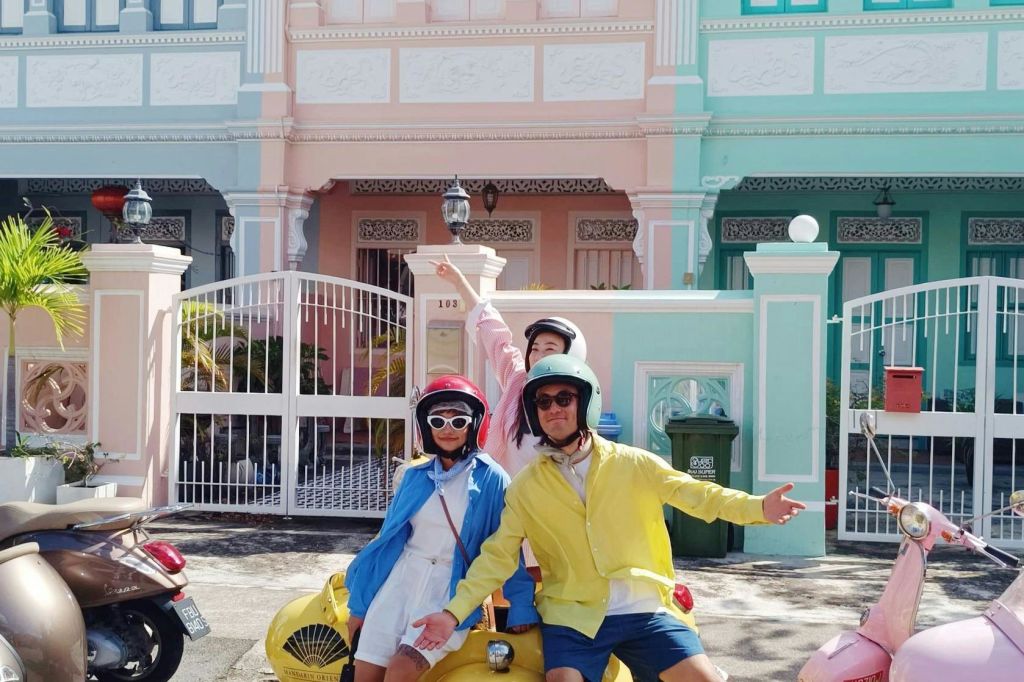 화창한 날, 파스텔 색상의 건물 앞에서 노란색 스쿠터를 타고 화려한 의상을 입은 세 사람이 포즈를 취하고 있습니다.