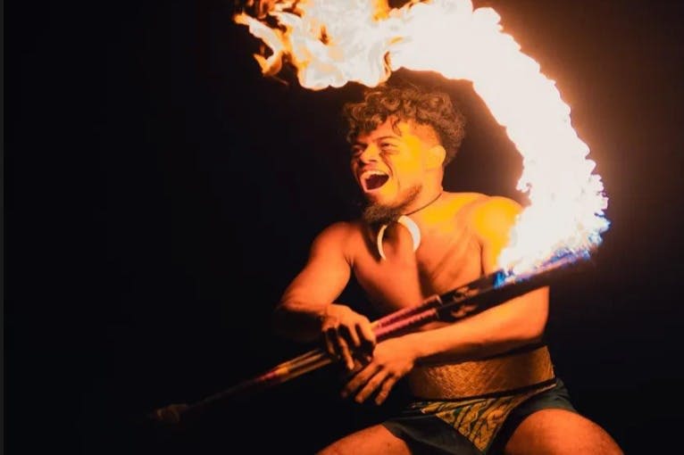 한 남성이 어두운 배경에 전통적인 복장을 한 채 불타는 막대기를 들고 공연을 합니다.