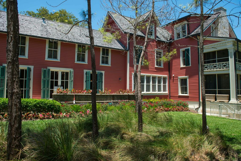 푸른 하늘 아래 잔디, 식물, 나무로 둘러싸인 녹색 셔터가 있는 커다란 빨간색 집입니다.