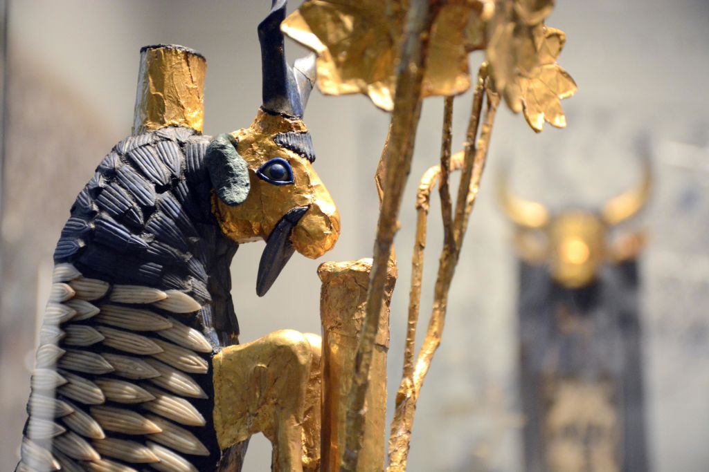 금빛 나무 옆에 검은색, 파란색, 녹색 디테일이 있는 염소를 묘사한 고대 황금 유물의 클로즈업.