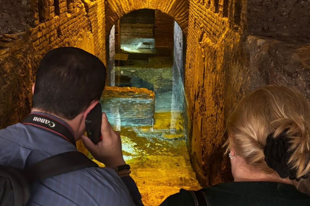 조명이 켜진 벽과 중앙 통로가 있는 고대 석조 및 벽돌 고고학 유적지를 관찰하는 두 사람.