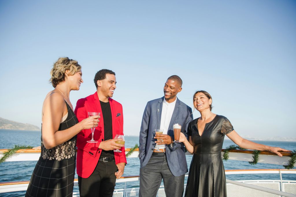 야외에서 배를 타고 서서 음료를 마시고 바다를 배경으로 미소를 짓는 네 명의 사람이 정장 차림을 하고 있습니다.