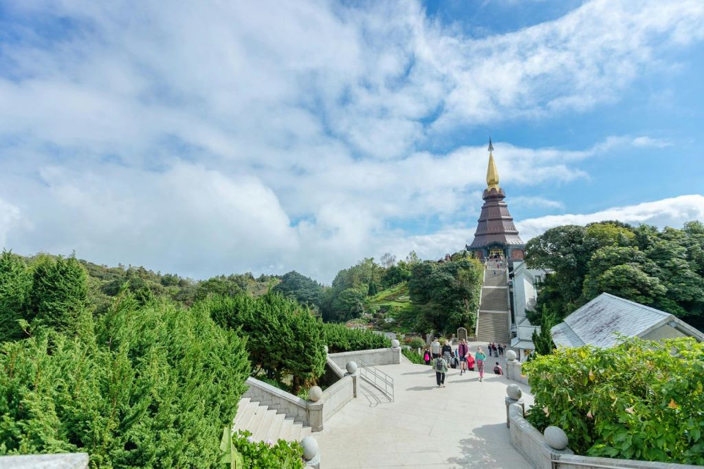 녹음으로 둘러싸인 황금 첨탑이 있고 부분적으로 흐린 하늘 아래 높이 솟은 탑을 향해 넓은 계단을 오르는 사람들.