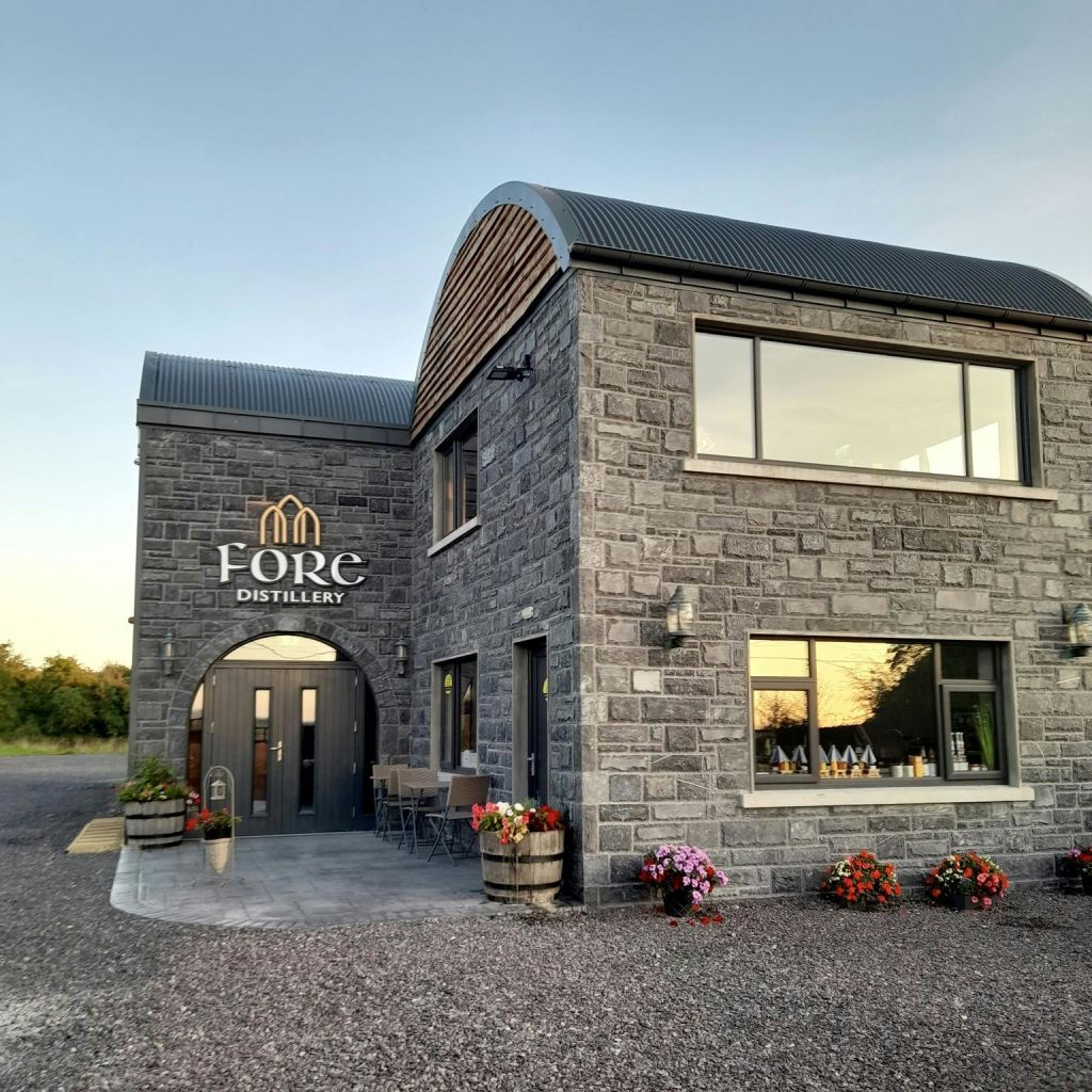 대형 창문, 야외 좌석, 화분, 자갈 통로가 있는 "Fore Distillery" 라고 표시된 석조 건물입니다.