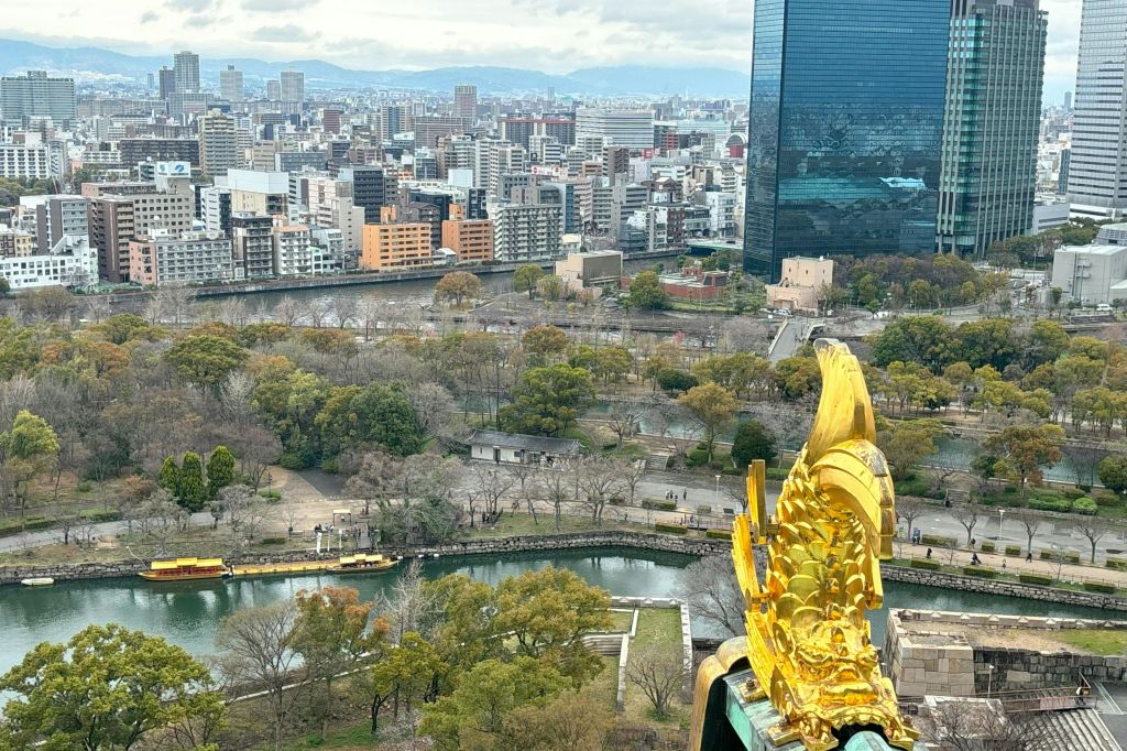 녹지 공원과 빽빽한 도시 건물이 어우러진 도시 경관으로, 황금 물고기 조각상이 전면에 눈에 띕니다.