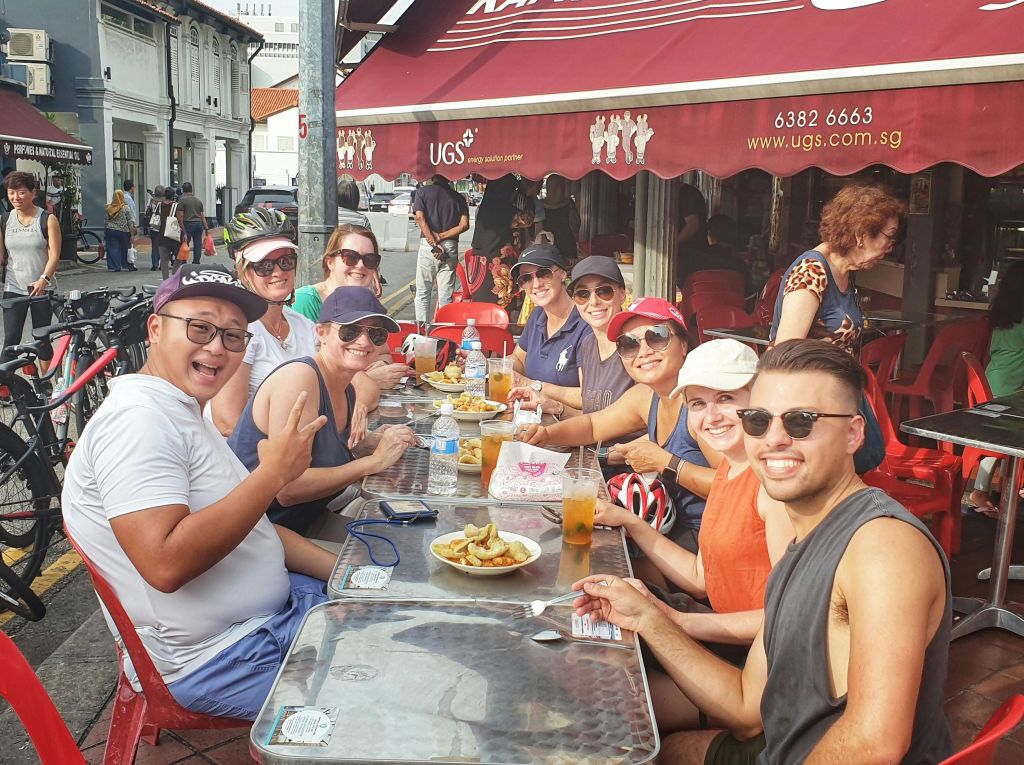 한 무리의 사람들이 야외 카페 테이블에 앉아 빨간색 캐노피 아래에서 음식과 음료를 즐기고 있습니다.