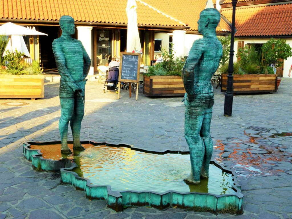 타일로 마감된 지붕이 있는 건물 근처의 마당에 얕은 물에 남성 두 금속 조각상이 서 있습니다.