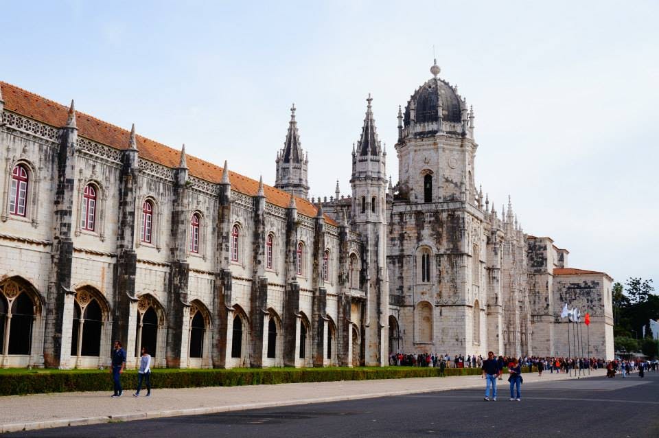 뾰족한 아치, 화려한 탑, 돔 구조물이 있는 역사적인 석조 건물. 사람들이 근처의 포장도로를 걷고 있습니다.