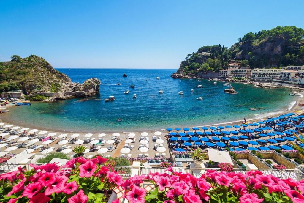 맑고 푸른 바다, 비치 파라솔, 물 속의 보트, 바위 절벽, 활기찬 분홍색 꽃이 눈앞에 펼쳐지는 해안 풍경.