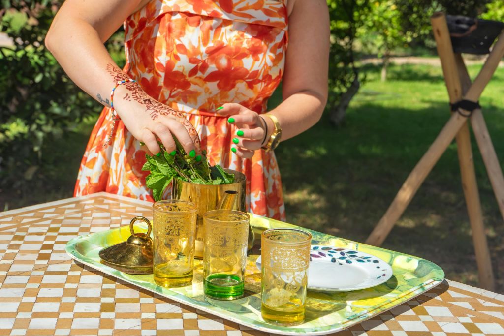 꽃무늬 드레스를 입은 사람은 야외에서 안경, 주전자, 접시가 있는 트레이에 민트 차를 준비합니다.
