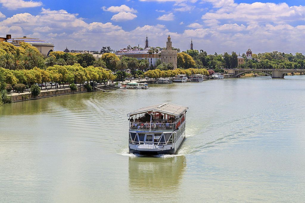 울창한 녹음과 유서 깊은 건물을 배경으로 부분적으로 흐린 하늘 아래에서 보트를 타고 강을 항해합니다.