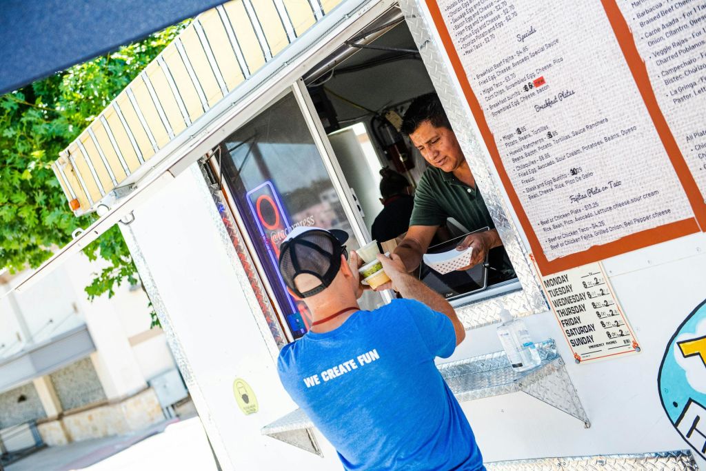 푸른색 셔츠를 입은 남성이 푸드트럭 안에서 판매자로부터 음식을 받고 있습니다. 메뉴는 트럭 측면에 표시됩니다.