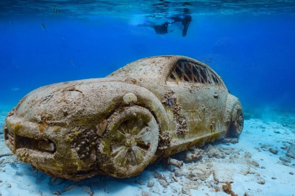 맑고 푸른 물 속에서 스쿠버 다이버가 헤엄치는 해초로 덮인 물에 잠긴 자동차를 보여주는 수중 장면.