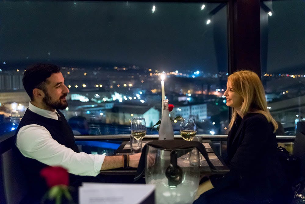 한 여성이 촛불이 켜진 저녁 식사에서 미소를 짓고 있으며, 밤에는 커다란 창문을 통해 도시 풍경을 감상할 수 있습니다. 테이블 위에 와인 두 잔이 놓여 있습니다.