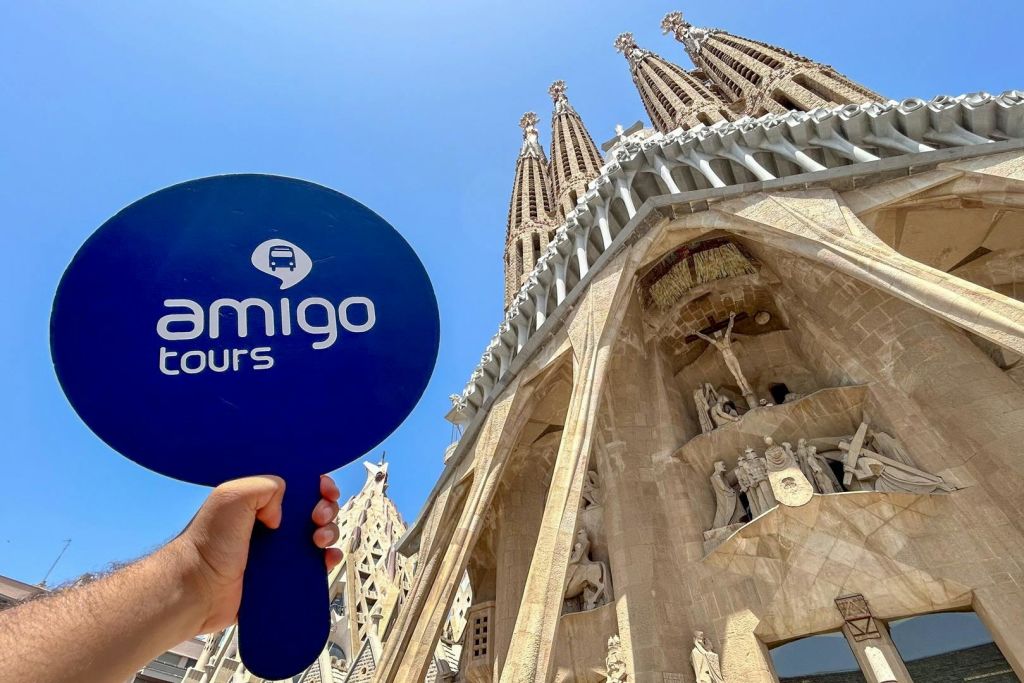 한 사람이 맑고 푸른 하늘 아래 사그라다 파밀리아 (Sagrada Familia) 앞에서 '아미고 투어 (Amigo Tours)' 라고 적힌 파란색 원형 표지판을 들고 있습니다.