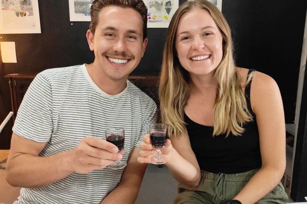 어두운 음료와 함께 작은 안경을 들고 미소를 짓고 있는 두 사람이 검은색 벽을 배경으로 실내에 앉아 있습니다.