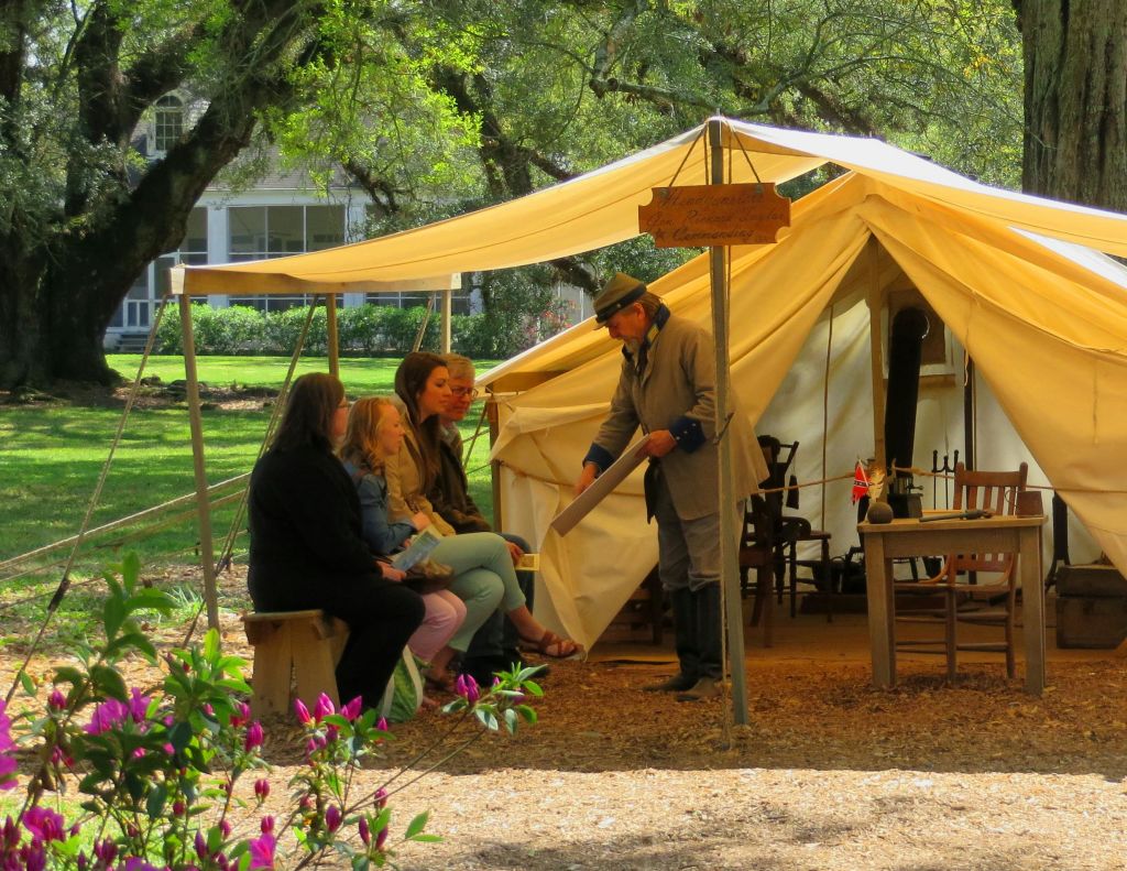 캔버스 텐트 앞 벤치에 네 명이 앉아 있고, 역사적인 복장을 한 남성이 이야기를 나누고 있습니다. 나무와 집이 배경입니다.