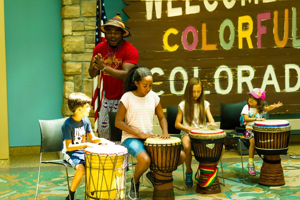 아이들이 "컬러풀 콜로라도에 오신 것을 환영합니다" 표지판 앞에서 강사와 함께 드럼을 연주합니다.