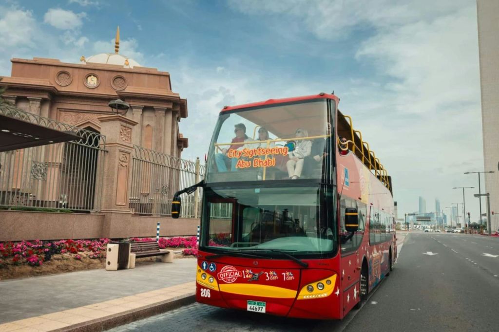 전면에 "도시 관광 아부다비" 가 있는 빨간색 2층 버스가 황금색 돔이 있는 화려한 건물을 지나갑니다.